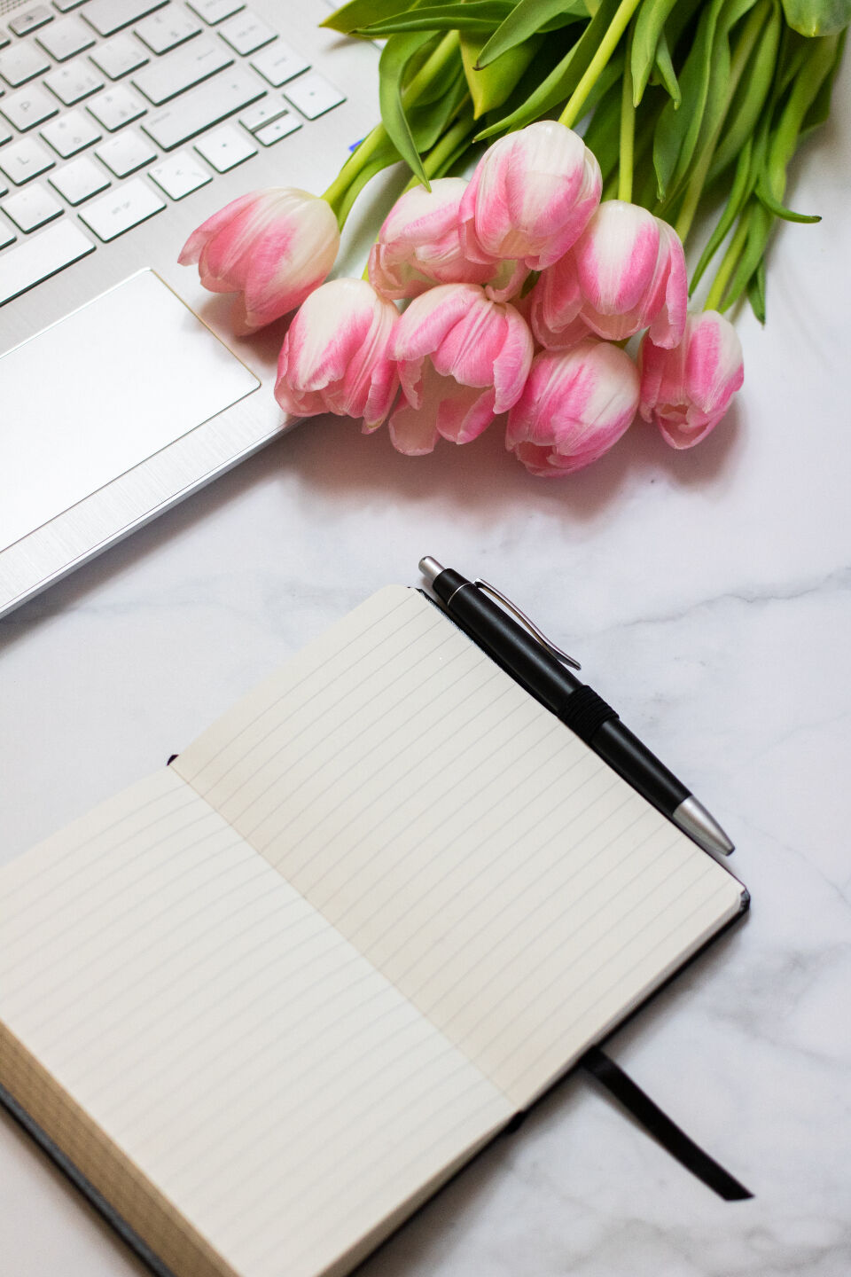 A laptop on a desk with flowers, a notebook and a pen